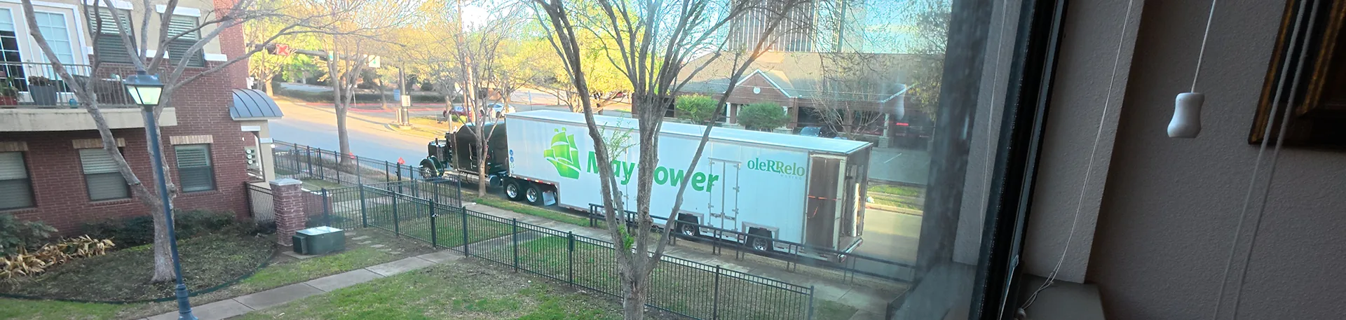 In-State Moving Day | Oler Relo Moving A Mayflower moving trailer parked on the street, seen through a home's window during an in-state move